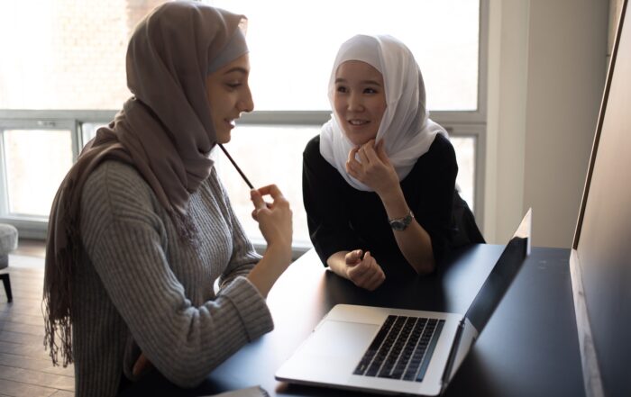 Positive multiethnic Muslim female students in traditional headscarves sitting at table and surfing netbook while preparing for lesson together in university