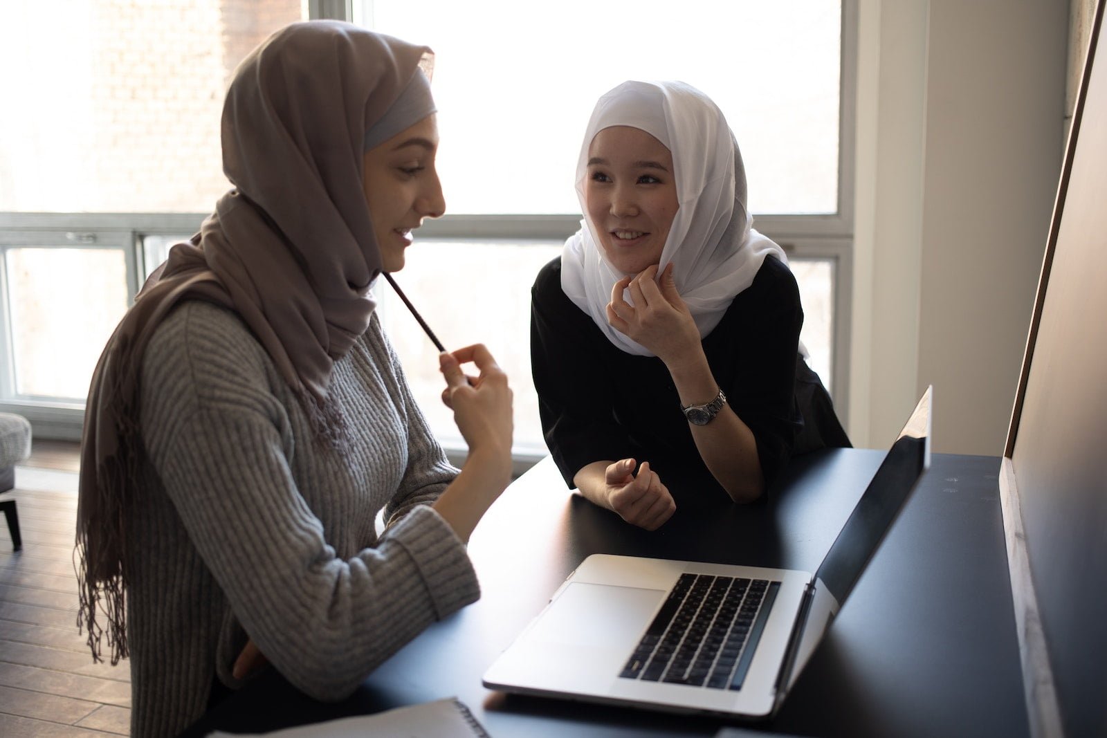 Positive multiethnic Muslim female students in traditional headscarves sitting at table and surfing netbook while preparing for lesson together in university